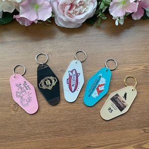 May include: Five colorful keychains with different designs. The keychains are shaped like motel room keys. The designs include a black keychain with a gold "JL" logo, a white keychain with a red, white, and blue airship with the word "AKRON" on it, a blue keychain with a red house and the text "Big Country Realty Group", a pink keychain with a black and white illustration of a person with a chain and the text "Big Jerry Tattoos", and a cream keychain with a black and white illustration of a bird and the text "Carlyle's 95".