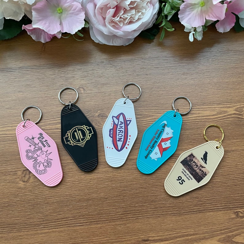 May include: Five colorful keychains with different designs. The keychains are shaped like motel room keys. The designs include a black keychain with a gold "JL" logo, a white keychain with a red, white, and blue airship with the word "AKRON" on it, a blue keychain with a red house and the text "Big Country Realty Group", a pink keychain with a black and white illustration of a person with a chain and the text "Big Jerry Tattoos", and a cream keychain with a black and white illustration of a bird and the text "Carlyle's 95".