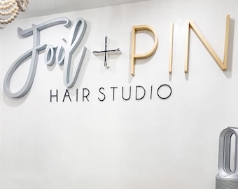 Könnte beinhalten: Eine weiße Wand mit einem goldenen und grauen Schild mit der Aufschrift "Foil + PIN HAIR STUDIO".