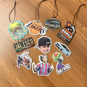 May include: Assortment of car air fresheners with various designs, including a basketball team logo, a car detailing company logo, a mobile mechanic logo, a cartoon crab, a state of Jalisco license plate, a child's face, a soccer player, and a cartoon character with a watermelon on its head.