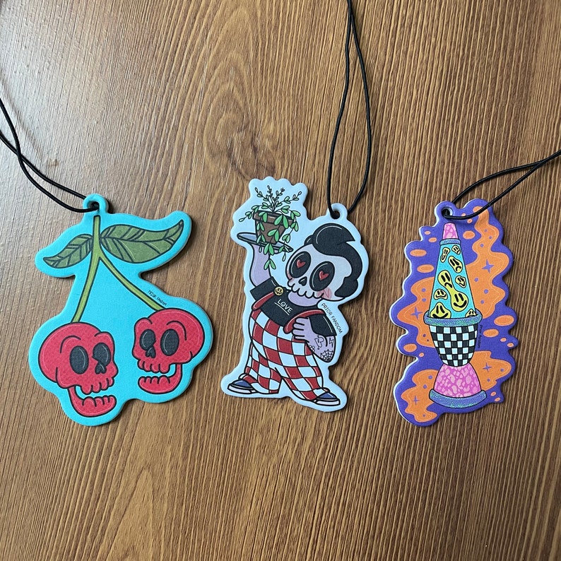 May include: Three air freshener car charms. One features a cherry with two skulls, another a cartoon character holding a plant, and the last a lava lamp with smiley faces.