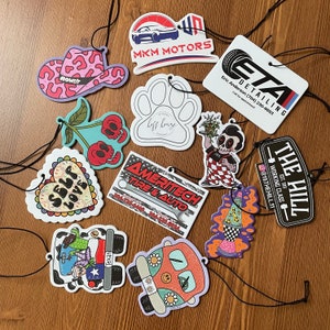 May include: A collection of ten colorful car air fresheners with various designs. The designs include a cowboy hat, a paw print, a cherry, a heart, a tire, a rocket, a van, and a logo for a car detailing business. The air fresheners are all on a wooden surface.