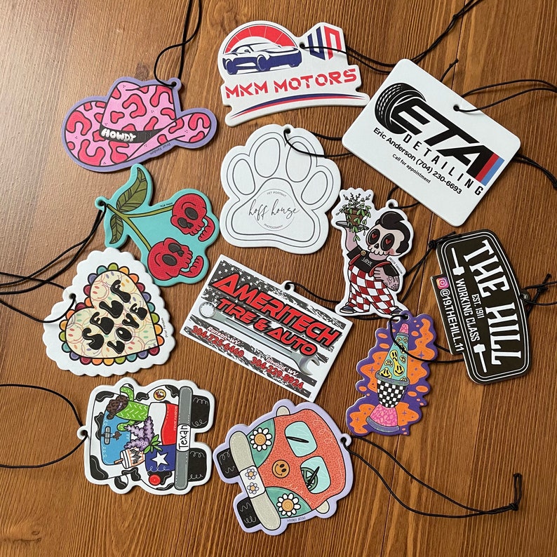May include: A collection of ten colorful car air fresheners with various designs. The designs include a cowboy hat, a paw print, a cherry, a heart, a tire, a rocket, a van, and a logo for a car detailing business. The air fresheners are all on a wooden surface.
