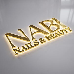 May include: A gold-colored sign that reads "NABI NAILS & BEAUTY" in a stylized font. The letters are raised and have a reflective surface, with a subtle glow emanating from behind. The sign is set against a neutral, light-colored background.