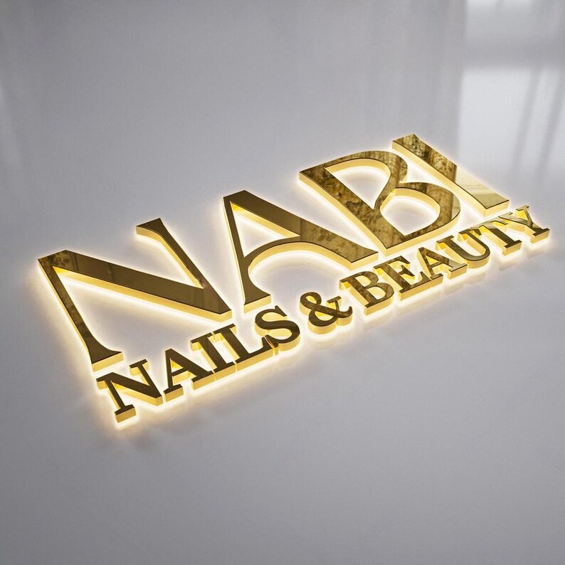 May include: A gold-colored sign that reads "NABI NAILS & BEAUTY" in a stylized font. The letters are raised and have a reflective surface, with a subtle glow emanating from behind. The sign is set against a neutral, light-colored background.
