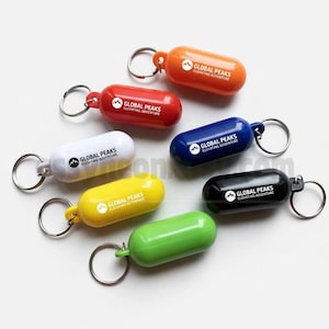 May include: A collection of colorful, capsule-shaped keychains in orange, red, white, blue, yellow, black, and green. Each keychain has a silver ring and the text "GLOBAL PEAKS ELEVATING ADVENTURE" printed on it.