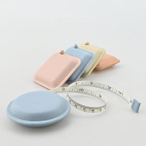 Op de afbeelding: Een verzameling pastelkleurige, vierkante meetlintetuis met een bijpassend blauw meetlint. Het meetlint is wit met zwarte markeringen en een blauwe punt. De etuis zijn in tinten roze, blauw, geel en perzik.