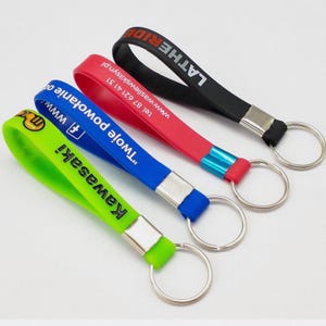 Könnte beinhalten: Vier farbige Armbänder mit Schlüsselringen. Die Armbänder sind grün, blau, rot und schwarz. Auf dem grünen Armband steht das Wort "Kawasaki". Auf dem blauen Armband stehen die Worte "Twoje powołanie". Auf dem roten Armband sind eine Telefonnummer und eine Website aufgedruckt. Auf dem schwarzen Armband steht das Wort "LATHERIDE".