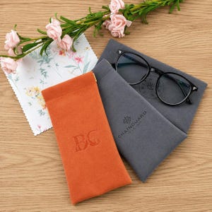 May include: An orange glasses case with the embossed initials "BC" next to a gray glasses case with the text "CHAINGUARD CLUB TRIP". Black-framed eyeglasses rest on the gray case. A floral print cloth and pink roses are also present.