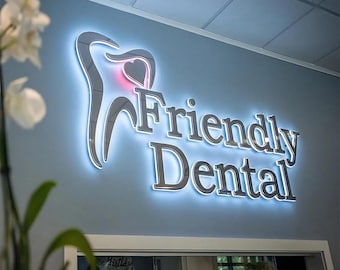 May include: A wall-mounted sign for a dental office. The sign features the words "Friendly Dental" in silver lettering, with a stylized tooth graphic and a heart shape. The sign is illuminated with a blue glow.