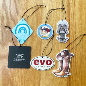 May include: Five different car air fresheners with various designs. The designs include a rainbow with the text "Be Their Voice", a superhero with the text "Superman", a skull with its hands over its eyes, a cowboy boot, and a logo with the text "evo HELMETS".