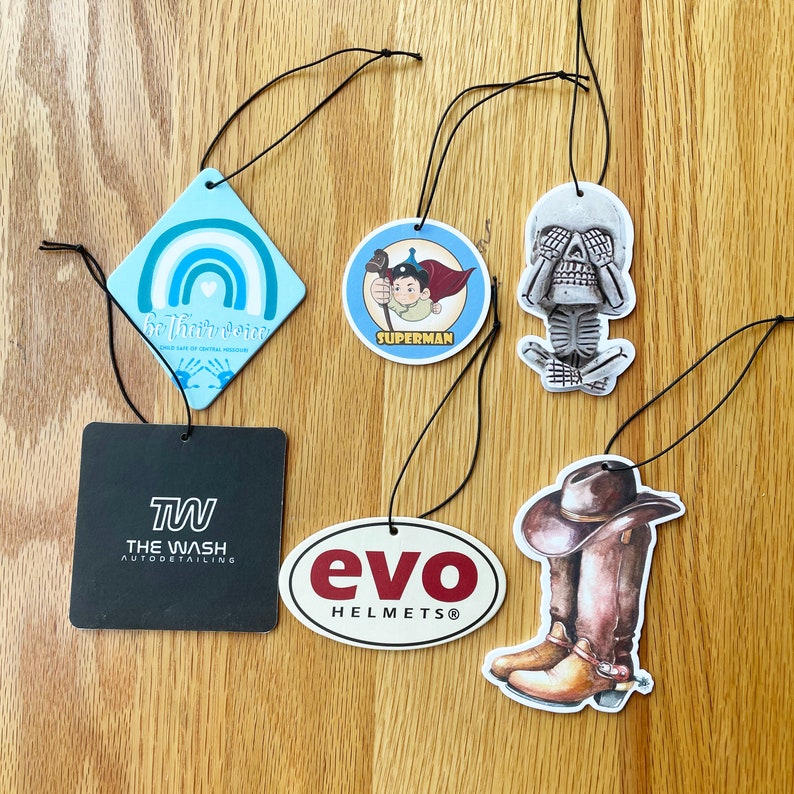 May include: Five different car air fresheners with various designs. The designs include a rainbow with the text "Be Their Voice", a superhero with the text "Superman", a skull with its hands over its eyes, a cowboy boot, and a logo with the text "evo HELMETS".