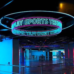 Puede incluir: Un letrero de neón circular suspendido del techo, con el texto PLAY SPORTS TREND en letras cian brillantes delineadas en rosa. El letrero está colocado dentro de un marco circular más grande, emitiendo un brillo vibrante en un espacio oscuro.