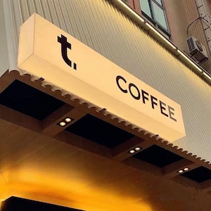 May include: Illuminated rectangular sign with the word "COFFEE" in a bold, black font. The sign also features a stylized black "t." symbol. The sign is mounted on a building facade with decorative trim and recessed lighting.
