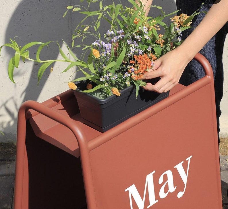 Puede incluir: Un letrero de metal en forma de A de color terracota con la palabra May impresa en letras blancas. Una jardinera rectangular negra se asienta en el borde superior, llena de follaje verde, peque&ntilde;as flores moradas y racimos de flores naranjas.