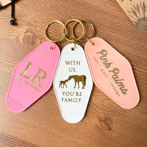 May include: Three pink and white keychains with gold lettering. The keychains are shaped like motel room keys. The first keychain says "LR local stay rentals". The second keychain says "With Us, You're Family" and has a silhouette of a horse and foal. The third keychain says "Pink Palms sun pool lounge".