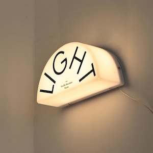 May include: A white, semi-circular wall light fixture with the word "LIGHT" in black lettering. The text "Vanilla Mountain house" is below the word "LIGHT". The fixture is mounted on a light gray wall.