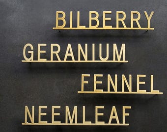 Könnte beinhalten: Vier messingfarbene Metallschilder mit den Wörtern "BILBERRY", "GERANIUM", "FENNEL" und "NEEMLEAF" auf dunklem, grauem Hintergrund. Jedes Wort ist in einer serifenlosen Schriftart und auf einem kleinen Regal angebracht.