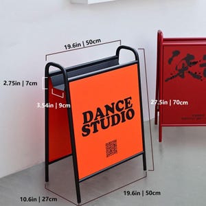 Puede incluir: Un letrero de metal negro en forma de A con un panel naranja que presenta el texto negro DANCE STUDIO y un c&oacute;digo QR. Las dimensiones son 50 cm de ancho, 70 cm de alto, 27 cm de profundidad, con una abertura de 7 cm y 9 cm de profundidad.