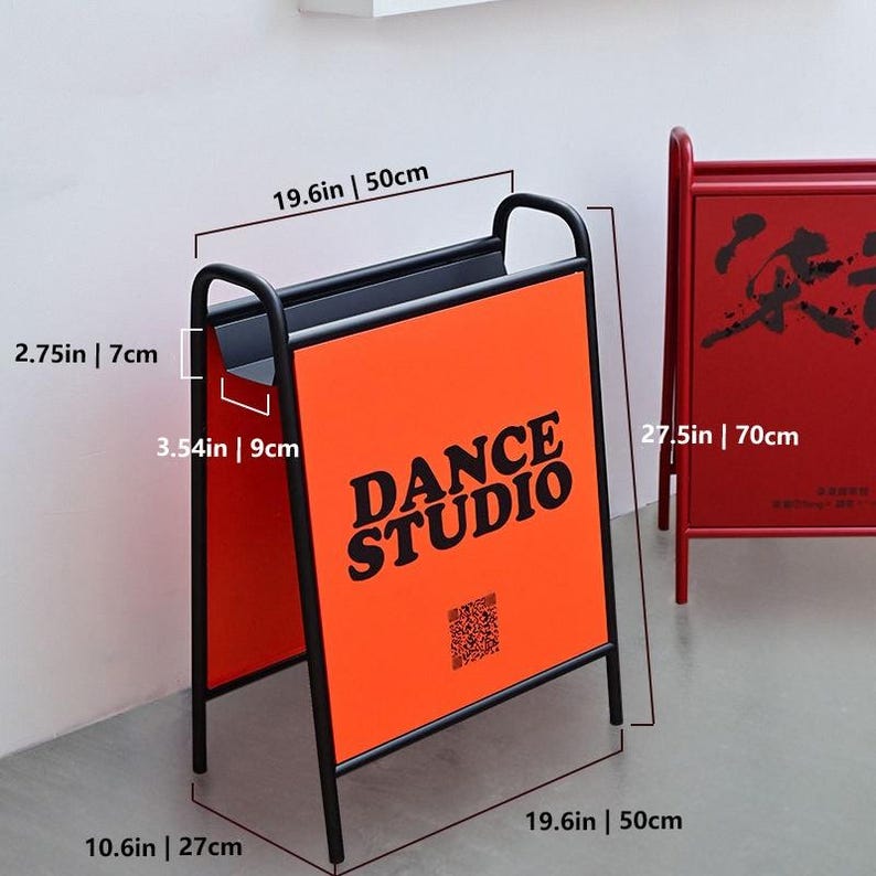 Puede incluir: Un letrero de metal negro en forma de A con un panel naranja que presenta el texto negro DANCE STUDIO y un c&oacute;digo QR. Las dimensiones son 50 cm de ancho, 70 cm de alto, 27 cm de profundidad, con una abertura de 7 cm y 9 cm de profundidad.