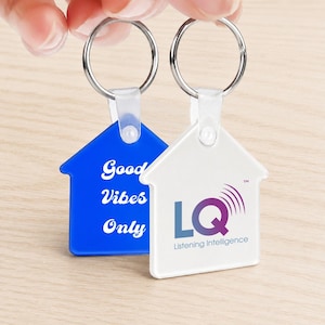 Peut inclure: Deux porte-clés en forme de maison. L'un est bleu avec les mots "Good Vibes Only" en blanc. L'autre est blanc avec un logo violet et bleu et le texte "LQ Listening Intelligence". Les deux ont des anneaux argentés.