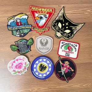 Könnte beinhalten: Eine Sammlung von neun gestickten Patches mit verschiedenen Designs, darunter eine schwarze Katze, ein roter Adler, eine Erdbeere, ein Herz und eine Fee. Die Patches sind alle in verschiedenen Größen und Formen und haben eine Vielzahl von Farben und Texturen.