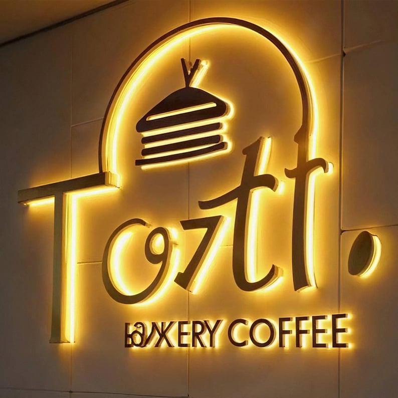 May include: Illuminated sign for a bakery and coffee shop. The sign features the name "Tozti" in a stylized font, with a graphic of a stack of pancakes above the name. The words "Bakery Coffee" are below the name.