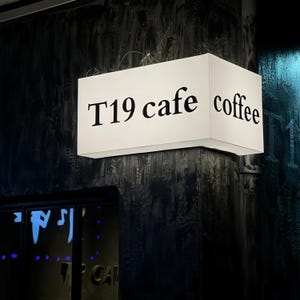 Puede incluir: Un letrero rectangular iluminado para una cafetería. El letrero es blanco con las palabras "T19 cafe coffee" en letras negras. El letrero está montado en una pared oscura y texturizada.
