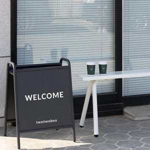 Puede incluir: Un letrero de acera negro en forma de A con el texto blanco WELCOME y twotwobox impreso en el frente. Junto al letrero hay un banco de metal blanco con dos vasos de caf&eacute; desechables de color verde oscuro con tapas blancas frente a una ventana.