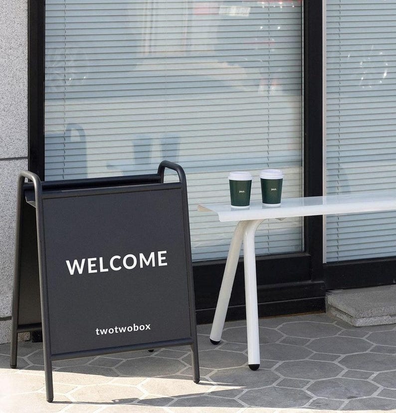Puede incluir: Un letrero de acera negro en forma de A con el texto blanco WELCOME y twotwobox impreso en el frente. Junto al letrero hay un banco de metal blanco con dos vasos de caf&eacute; desechables de color verde oscuro con tapas blancas frente a una ventana.