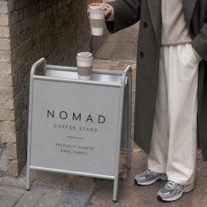 Puede incluir: Un letrero de metal gris claro en forma de A con el texto NOMAD COFFEE STAND SPECIALTY COFFEE MADE SIMPLE. Dos tazas de caf&eacute; reutilizables blancas con fundas texturizadas de color beige est&aacute;n colocadas en el borde superior del letrero.