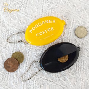 May include: Two oval coin purses, one yellow and one black, with attached keychains. The yellow purse has the text "PONGANES COFFEE" and a website address. Several coins are scattered around the purses.