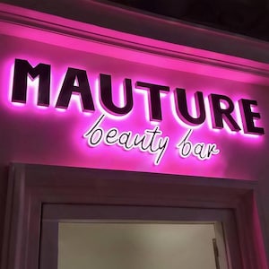 May include: A sign for a beauty bar with the word "MAUTURE" in large, dark letters, illuminated with a pink glow. Below, the words "beauty bar" are written in a cursive font, also with a pink glow.