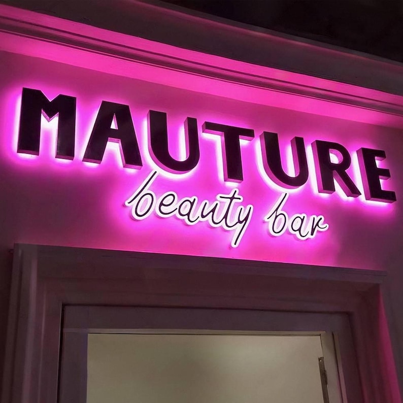 May include: A sign for a beauty bar with the word "MAUTURE" in large, dark letters, illuminated with a pink glow. Below, the words "beauty bar" are written in a cursive font, also with a pink glow.