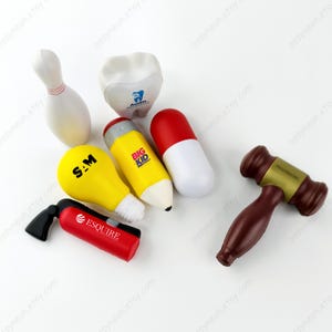 May include: A collection of stress-relieving toys in various shapes and colors. Includes a white bowling pin, a tooth with a blue logo, a yellow lightbulb with "S.A.M" printed on it, a yellow pencil with "BIG KID" printed on it, a red and white pill, a red fire extinguisher with "ESQUIRE" printed on it, and a brown gavel.