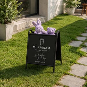 Puede incluir: Un letrero de acera negro en forma de A con un icono de taza de caf&eacute; blanca, el texto MILLIGRAM COFFEE STAND, la frase good coffee, good day y el usuario @milligram.coffee, con un conejo y un oso de peluche de color violeta claro.