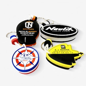 Könnte beinhalten: Eine Sammlung von vier Schlüsselanhängern mit verschiedenen Designs. Einer ist schwarz mit weißem Text und Logo, ein anderer gelb und schwarz mit Text und Logo, und zwei sind rund mit nautischen Themen. Jeder Schlüsselanhänger hat einen Metallring.