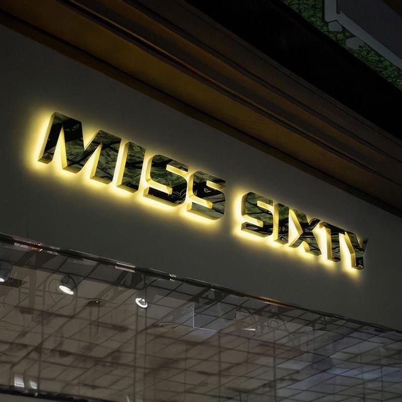 May include: Illuminated "MISS SIXTY" sign in a store front. The letters are metallic with a reflective surface, and are lit from behind with a warm, yellow glow. The sign is mounted on a neutral-colored wall, with a glass storefront below.