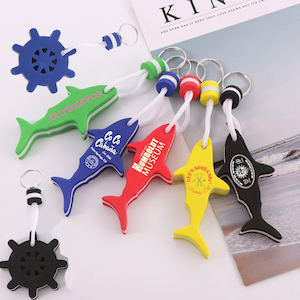 Puede incluir: Una colección de llaveros con forma de tiburón de colores, y un llavero azul con forma de timón. Los llaveros de tiburón son de varios colores, incluyendo verde, azul, rojo, amarillo y negro, con cordones blancos y anillas metálicas. Algunos tienen texto.