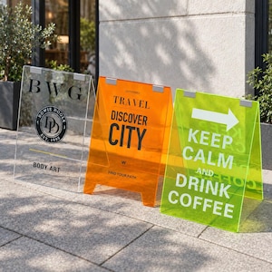 Puede incluir: Tres letreros tipo caballete de acrílico transparente en colores claro, naranja y verde lima. El letrero claro tiene texto negro que dice BWG, DOWIE DOOLE y BODY ART. El letrero naranja muestra texto negro que dice TRAVEL DISCOVER CITY y FIND YOUR PATH. El letrero verde muestra texto blanco que dice KEEP CALM AND DRINK COFFEE con una flecha.