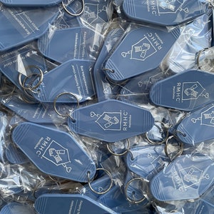 May include: A pile of blue keychains with the text "RMHC Manitoba" and the slogan "Keeping families close" printed on them. The keychains are shaped like motel keys and have a silver key ring attached.
