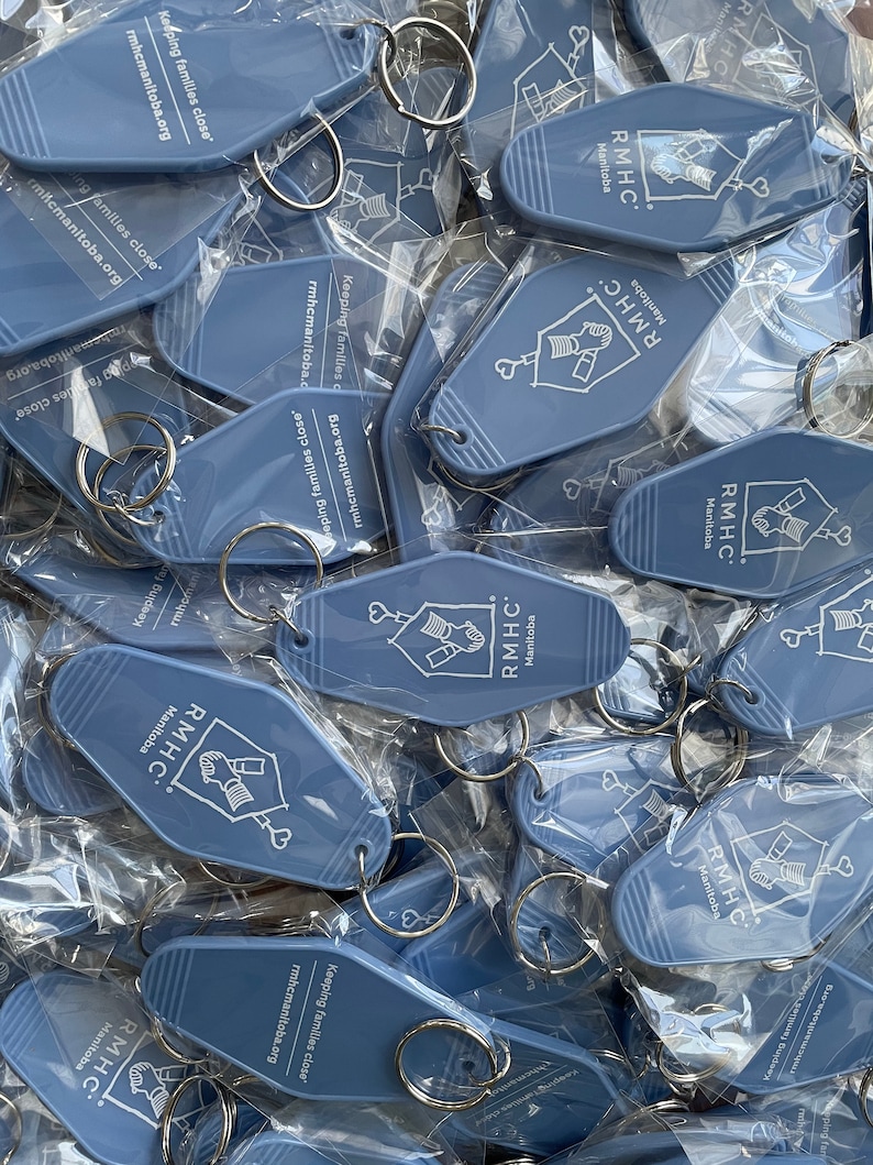 May include: A pile of blue keychains with the text "RMHC Manitoba" and the slogan "Keeping families close" printed on them. The keychains are shaped like motel keys and have a silver key ring attached.