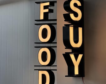 May include: Illuminated sign in black, spelling out "FOOD" and "SUY". The letters are lit with a warm, yellow glow. The sign is mounted on a wall, with a red wire visible.