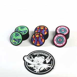 May include: A selection of circular, black rubber patches with vibrant designs. Some patches feature geometric patterns in green, purple, and orange. One patch displays a white and black illustration with the text "TENGUMASK".