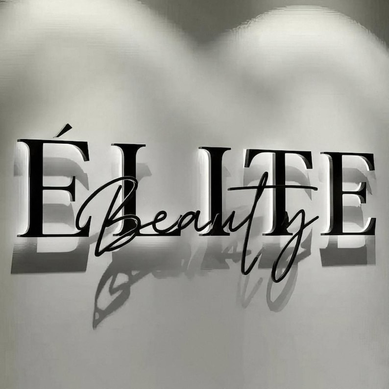 May include: Black illuminated sign on a white wall. The sign reads "&Eacute;LITE Beauty" in a combination of bold, capital letters and elegant cursive script. The letters cast shadows, creating a three-dimensional effect. The overall aesthetic is modern and sophisticated.