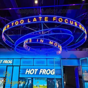 May include: A blue neon sign with the text "Too Late Focus On" in yellow letters. The sign is hanging from the ceiling of a shop with the name "Hot Frog" in white letters on a blue background.