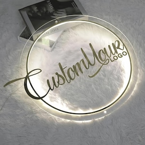 Può includere: Un cartello rotondo in acrilico trasparente con una scritta dorata che recita "Custom Your Logo". Il cartello è illuminato da un anello di luce LED bianco.