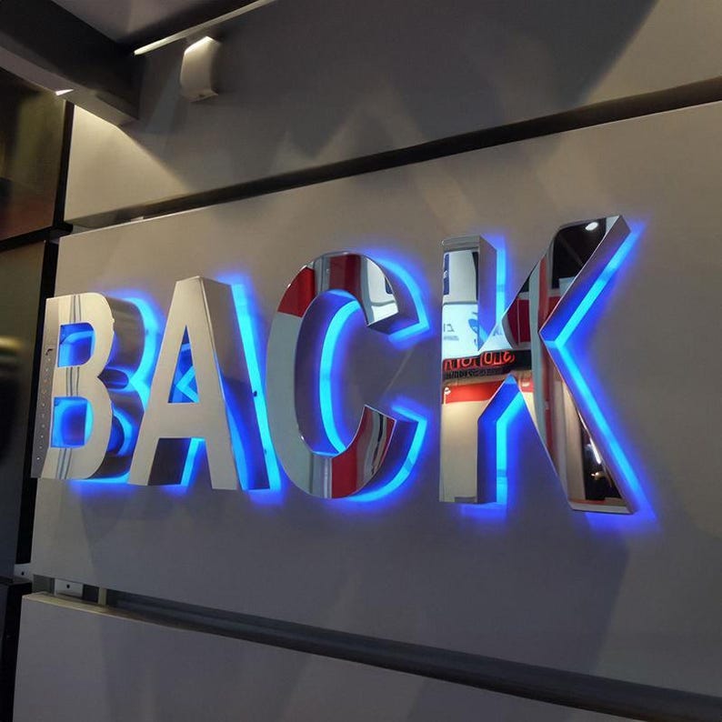 May include: A wall-mounted sign that spells out the word "BACK" in large, reflective, silver-colored letters. The letters are outlined with a bright blue neon glow, creating a striking visual effect. The sign is mounted on a light gray wall.