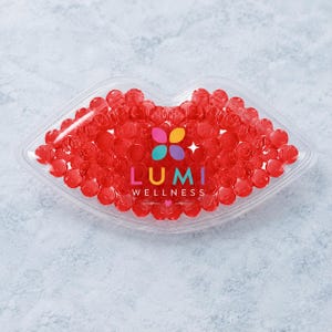 Puede incluir: Un recipiente transparente con forma de labios lleno de pequeñas cuentas rojas redondas y translúcidas. El centro del recipiente presenta un logotipo floral multicolor sobre el texto "LUMI WELLNESS" impreso en blanco, con un pequeño corazón debajo.