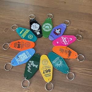 May include: A collection of ten colorful keychains with various designs. The keychains are shaped like motel keys and feature different text and graphics, including the words "The Clubhouse Collective", "Armed & Courageous", "The Adler", "John", "Motel Sunday 867", and "Room 152".